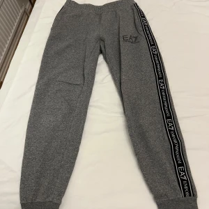 Grå EA7 Emporio Armani mjukisbyxor - Grå mjukisbyxor från EA7 Emporio Armani med svart logoband längs sidorna. Byxorna har elastisk midja, muddar vid benslut och är tillverkade i en mjuk bomullsblandning. Perfekta för chill eller träning, med snygg och sportig look.