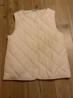Beige quiltad dunväst barn 146/152 - Säljer en beige quiltad dunväst för barn i storlek 146/152. Västen har tryckknappar framtill och en enkel, stilren design utan krage. Perfekt att slänga över en hoodie eller tröja för extra värme. Aldrig använd 