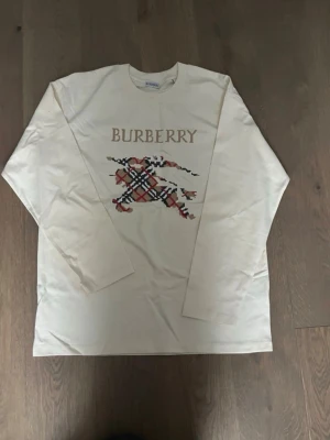 Burberry tröja  - Snygg långärmad tröja från Burberry i ljus beige bomull. Framsidan har ett stort broderat Burberry-tryck och klassiskt rutig mönster i rött, svart och beige. Perfekt för dig som gillar stilrena och exklusiva plagg med en twist. Knappt använd säljes pga av att det bara sitter i garderob… Storlek M