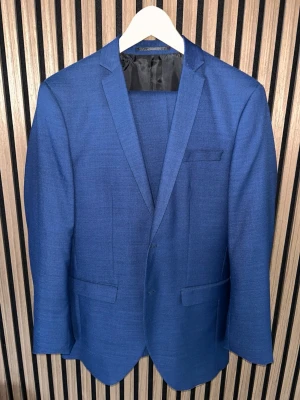 Blå kavaj set med kostymbyxor från Jack & Jones - Snygg marinblå kavaj från Jack & Jones i super slim passform. Kavajen har klassisk krage, tre fickor fram och fyra svarta knappar vid ärmslutet. Materialet är troligtvis polyester och kostymen  har en stilren look som passar vid lite finare tillställningar. Hör av er vid intresse/ frågor!