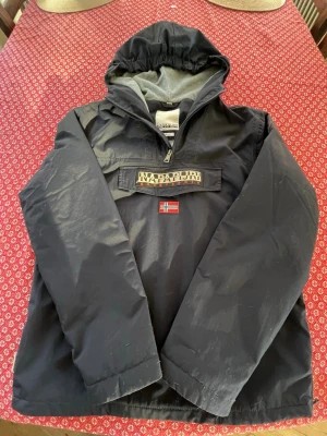 Svart vindjacka från Napapijri - Säljer en svart vindjacka från Napapijri med huva och stor ficka framtill med dragkedja och kardborre. Jackan har Napapijri-logga och norsk flagga på bröstet. Insidan är fodrad med mjukt grått tyg. Perfekt för kyliga dagar.ny pris 1799 mitt 799,priset är inte hugget i sten 