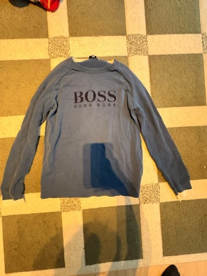 Blå tröja från Hugo Boss - Säljer en blå stickad tröja från Hugo Boss med stort BOSS-tryck på bröstet.👌Tröjan har rund halsringning, långa ärmar och ribbade muddar med vit detalj längst ner på ärmarna. Perfekt för dig som gillar klassisk och stilren design. Tveka inte om att skriva om pris förslag!😄