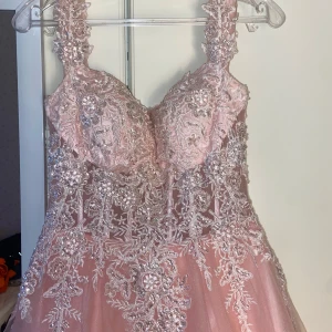 Rosa långklänning med spets och paljetter - Drömmig rosa långklänning med vackra spetsdetaljer och glittrande paljetter över hela livet. Klänningen har breda axelband, markerad midja och en voluminös kjol i flera lager tyll. Perfekt för bal eller quinceañera. Snyggt blommönster och lyxig känsla. Har bara använts en gång. 