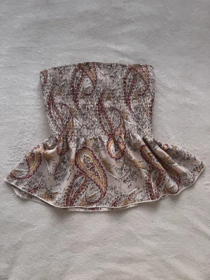 Beige paisleymönstrad linnetopp - Supersöt linnetopp i beige med paisleymönster i rött, gult och brunt. Smockad upptill för en tight passform över bysten och volang nedtill som ger en flowy känsla. Tunna axelband med spetsdetalj.