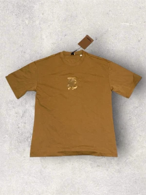 Beige Burberry t-shirt med logga - Snygg beige t-shirt från Burberry med guldfärgad logga på bröstet. Klassisk rund halsringning och korta ärmar. Tillverkad i mjuk bomull som känns skön mot huden. Perfekt för dig som vill ha en clean look med exklusiv touch.