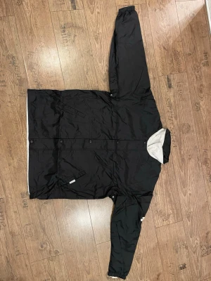 Svart Nike vindjacka med vita detaljer - Säljer en svart vindjacka från Nike med vita bokstäver på ena ärmen och vit insida. Jackan har knappstängning framtill och sidofickor. Knappt använd och inga skador