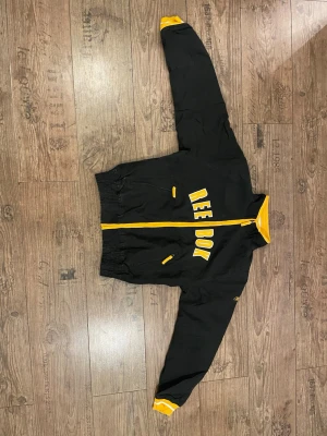 Svart och gul bomberjacka från Reebok - Säljer en svart bomberjacka från Reebok med gula detaljer och broderad text på bröstet. Jackan har dragkedja framtill, ribbade muddar och krage i gult samt siffran 56 på ryggen. Storlek XS. Jackan är använd en del.