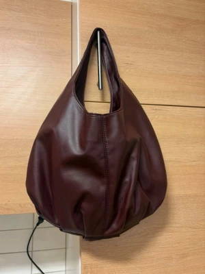 Vinröd axelväska från Bik Bok - Säljer en vinröd axelväska från Bik Bok i mjukt skinnimitation. Väskan har en rundad, slouchy form och stängs med magnetknapp. Insidan har en praktisk innerficka med dragkedja. Perfekt storlek för vardagens essentials.