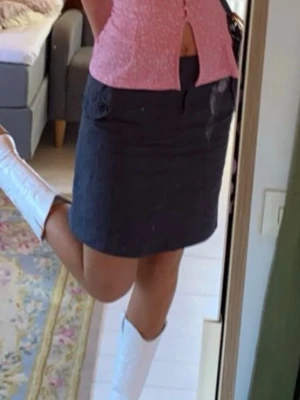 Svart kortkjol från Lindex - Säljer en svart kortkjol från Lindex. Kjolen har tunna vita ränder, två fickor framtill med knappdetaljer och stängs med dragkedja och någon mer grej:) Inte stretch i tyget.