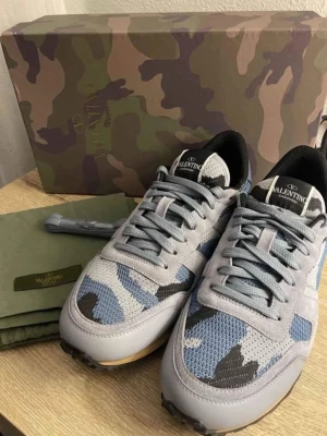 Valentino Garavani Rockrunner camo - Säljer ett par Valentino Garavani Rockrunner sneakers i grått, blått och svart med camouflage-mönster. Skorna har detaljer i mocka och mesh, samt coola nitar på hälen. Klassisk snörning och beige sula ger en unik look. Perfekta för dig som vill sticka ut.
