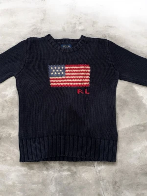 Mörkblå stickad tröja Polo Ralph Lauren - Mörkblå stickad tröja från Polo Ralph Lauren med amerikansk flagga broderad på bröstet och RL-bokstäver i rött. Tröjan har rund hals, ribbade muddar och är långärmad. Perfekt för kyliga dagar och ger en klassisk preppy look.