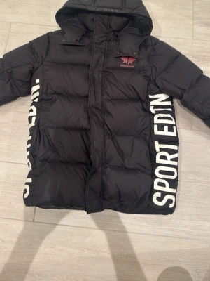 Svart pufferjacka från Dsquared2 - Säljer en svart pufferjacka från Dsquared2 med stor huva och feta vita SPORT EDTN-tryck längs sidorna. Jackan har dragkedja framtill och är riktigt vadderad för max värme. Perfekt för dig som vill ha en streetig och cool vinterlook.