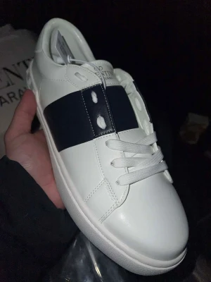 Valentino Garavani Open sneakers vit/svart - Säljer ett par vita Valentino Garavani Open sneakers med bred svart läderdetalj över mitten. Klassisk rund tå, vita snören och platt sula. Skorna är tillverkade i skinn och har en stilren, lyxig look som passar till det mesta.