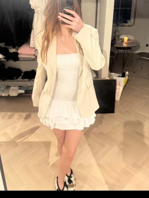 Vit axelbandslös volangklänning - Supersöt vit axelbandslös klänning med volanger nertill. Klänningen är kort och har en figurnära passform upptill med två lager volang i kjolen. Perfekt för sommarens alla tillfällen och lätt att styla med kavaj eller sneakers.