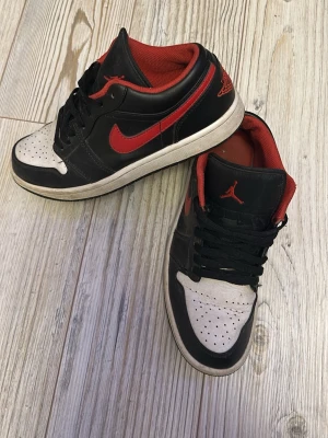 Nike Air Jordan 1 Low svart/röd - Säljer ett par Nike Air Jordan 1 Low sneakers i svart och rött med vita detaljer. Skorna har klassisk röd swoosh, svart skinnöverdel, perforerad vit tåbox och röd vadderad insida. Ikonisk Air Jordan-logga broderad på hälen och Jumpman på plösen. Perfekta för dig som gillar streetwear.