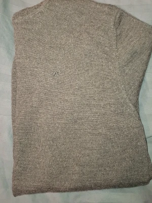 Grå stickad tröja från J.Lindeberg - Säljer en stilren grå stickad tröja från J.Lindeberg i mjuk och tunn stickad ull. Tröjan har hög krage, diskret logga broderad på bröstet och lång ärm. 