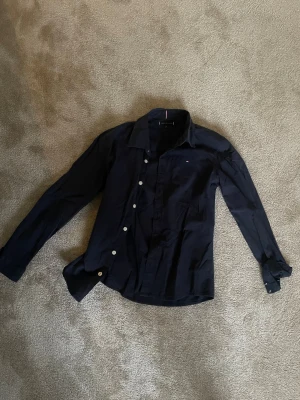 Mörkblå skjorta Tommy Hilfiger - Snygg mörkblå skjorta från Tommy Hilfiger med klassisk krage och vita knappar. Skjortan har lång ärm och en liten logga på bröstet. Tillverkad i mjuk bomull som känns skön mot huden. Perfekt för dig som vill ha en stilren och enkel look.