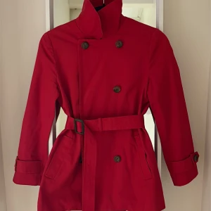 Röd trenchcoat från Zooc med bälte - Snygg röd trenchcoat från Zooc med klassisk dubbelknäppning, bälte i midjan och rutigt foder. Jackan har långa ärmar med spänne vid mudden och stilren krage. Perfekt för dig som vill sticka ut med färg och stil.