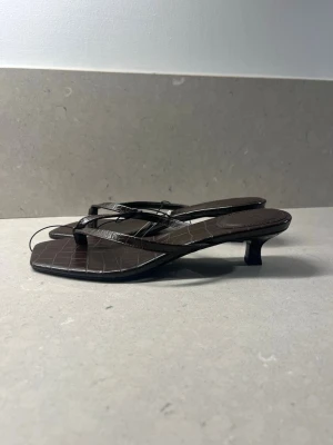Bruna sandaler med låg klack - Säljer ett par bruna sandaler med fyrkantig tå och låg klack. De har tunna remmar och ett croco-präglat mönster som ger en trendig look. Perfekta till sommaren och lätta att matcha med olika outfits.