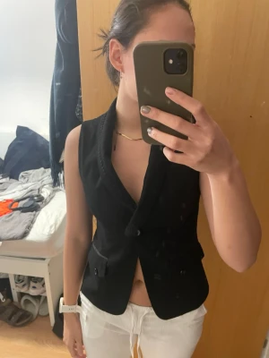 Svart kostymväst från Cortefiel - Snygg svart väst från Cortefiel med klassisk krage och knäppning framtill. Västen har två fickor med lock och knappar samt dekorativt band längs kragen.  Kan bäras som den är till fest eller med vanlig t shirt eller tight långärmad