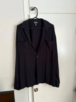 Svart overshirt från H&M - Overshirt i svart från H&M med två bröstfickor och knäppning framtill. Skjortan har en avslappnad passform, klassisk krage och är tillverkad i ett mjukt, ribbat material. Perfekt att slänga över en t-shirt för en chill look.