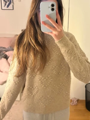 Beige stickad tröja med hjärtmönster - Mysig beige stickad tröja med upphöjt hjärtmönster över hela plagget. Tröjan har rund hals, ribbade muddar och lång ärm. 