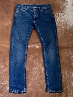 Blå jeans från Nudie Jeans Co W30 L32 - Säljer ett par klassiska blå jeans från Nudie Jeans Co i storlek W30 L32 Lean Dean. Jeansen har raka ben, fem fickor och kontrastsömmar samt Nudie-logga på bakfickorna. Tillverkade i slitstarkt bomullstyg med snygg tvättad look.