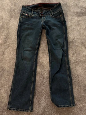 Lowrise jeans från Tommy Hilfiger - Snygga mörkblå jeans från Tommy Hilfiger med bootcut och femficksmodell. Byxorna har dubbla knappar i midjan, röda sömmar och broderad logga bak. De är i väldigt bra skick och inga synliga defekter💞💞 de passar en S/M