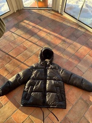 Svart pufferjacka med huva - Säljer en svart pufferjacka med huva och dragkedja framtill. Jackan har snörning i nederkant och stora fickor på sidorna. Perfekt för kalla dagar och riktigt skön att bära. Materialet är glansigt och känns lätt men ändå varmt. Den typiska hmjackan. Hör av dig vid frågor eller pris!
