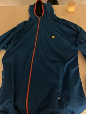 Blå vindjacka från Lyle & Scott - Säljer en blå vindjacka från Lyle & Scott med huva och hel orange dragkedja. Jackan har en gul logga på bröstet och svart meshfoder på insidan. Perfekt för dig som vill ha en sportig och stilren look.