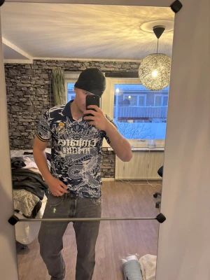 Real Madrid x Adidas blå draktröja - Säljer en blå och vit Real Madrid t-shirt från Adidas med fet drakmönster över hela tröjan och klubbmärke på bröstet. Klassisk krage och gula detaljer på ärmsluten. Perfekt för dig som vill sticka ut på läktaren eller i stan.