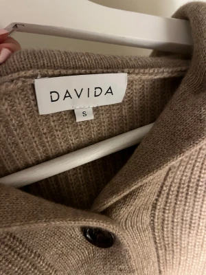 Beige stickad kofta från Davida - Supermysig beige stickad kofta från Davida i storlek S. Koftan har ribbstickat material, stora svarta/ bruna knappar framtill och en bred krage. Väldigt mysig, endast använd ca 2 gånger så inga noppor.