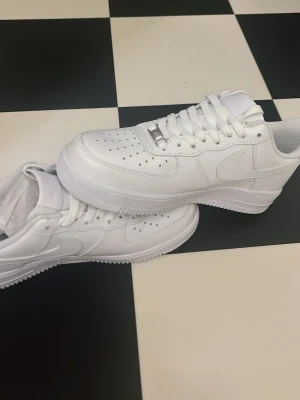 Nike Air Force 1 - Nike Air Force 1 sneakers i helvit färg med klassisk låg siluett. Skorna har snörning, perforerad tå för ventilation och rund tå. Tillverkade i skinn med platt sula och ikonisk Nike-logga på sidan och hälen. Perfekta för dig som gillar clean och stilren look. Helt oanvända! Tveka inte på att höra av er 