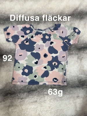 Blommig t-shirt i pastellfärger - Söt t-shirt med blommigt mönster i ljusrosa, blått, lila och grönt. Kortärmad modell med mjuk och bekväm känsla, perfekt för varma dagar. 🌸Diffusa fläckar. Storlek 92