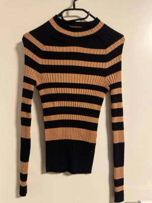 Randig ribbad tröja i svart och beige från Zara  - Säljer en tight, ribbad tröja med svarta och beigea ränder. Tröjan har lång ärm och en högre rund halsringning. Perfekt för dig som gillar en slim och figurnära passform med trendiga ränder.