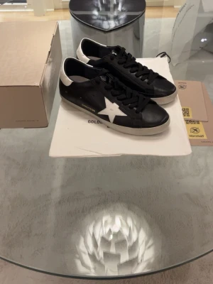 Svarta Golden Goose sneakers med vit stjärna - Golden Goose sneakers i svart skinn med vit stjärndetalj på sidan och vit häl. Skorna har svarta snören, rund tå och platt sula. GGDB/SSTAR-logga i guld på sidan. Snygg och stilren design som sticker ut med den ikoniska stjärnan.
