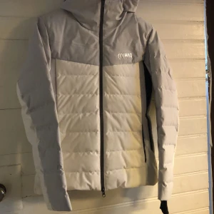 Ljusgrå dunjacka från Colmar - Snygg ljusgrå dunjacka från Colmar med huva och raka linjer. Jackan har svart dragkedja framtill och diskret Colmar-logga på bröstet. Fodrad insida och quiltad design för extra värme. Perfekt för kalla dagar och stilren look i skidbacken . Helt oanvänd .