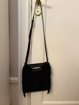Svart maje  -  en svart axelväska i mocka med fransar på sidorna och ett coolt handtag i toppen. Väskan har en lång axelrem och är perfekt för dig som vill ha en stilren men ändå unik accessoar. Insidan är rymlig och enkel utan extra fack.