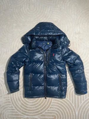 Mörkblå pufferjacka från Polo Ralph Lauren - Säljer en mörkblå pufferjacka från Polo Ralph Lauren i storlek XS. Jackan har huva, två dragkedjeförsedda fickor och en röd broderad logga på bröstet. Tillverkad i glansig polyester och stängs med dragkedja framtill. Perfekt för kalla dagar.