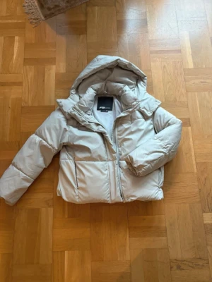 Beige pufferjacka från New Yorker - Säljer en beige pufferjacka från New Yorker med huva och dragkedja. Jackan har en puffig passform och är vadderad för extra värme. Fodrad insida och stora fickor framtill. Storlek xs