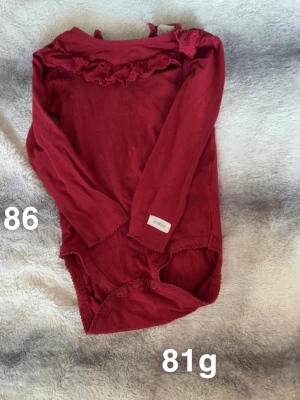 Röd body med volang och spets - Söt röd långärmad body från Newbie med volang och spetsdetaljer vid halsen. Knäppning i grenen för enkel påklädning. Perfekt för dig som gillar fina detaljer och en klassisk look.🌸 Fint använt skick. Storlek 86