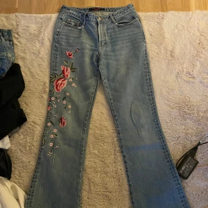 Blå broderade jeans från CFLine - Snygga ljusblå jeans från CFLine med broderade blommor i rosa och grönt längs ena benet och på bakfickan. Jeansen har klassisk femficksdesign och raka ben med lätt utsvängd passform. Perfekta för dig som vill sticka ut med detaljer!