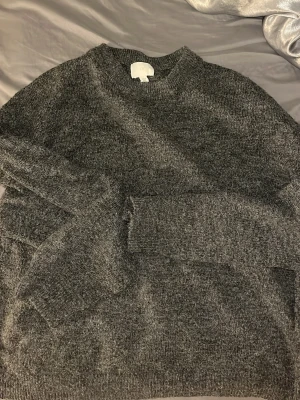 Mörkgrå stickad tröja från H&M - Mysig mörkgrå stickad tröja från H&M med rund halsringning och långa ärmar. Perfekt för kyliga dagar och enkel att matcha med olika outfits. Tröjan har en klassisk passform och mjukt material som känns skönt mot huden.