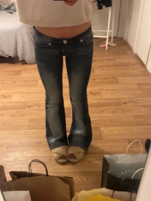 Lågmidjade bootcut jeans - Säljer ett par blå lågmidjade bootcut jeans med slitningar och klassisk femficksdesign. Jeansen har en snygg tvättad look med vinröda detaljer och är tillverkade i jeansmaterial. Perfekta för dig som gillar retrostil och vill ha ett par riktigt schyssta jeans.