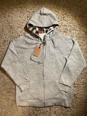 ljusgrå hoodie från Burberry  - Snygg ljusgrå hoodie från Burberry med klassiskt rutigt foder i huvan, broderad logga på bröstet och hel dragkedja. Tröjan har snörning i huvan och praktiska fickor framtill. Perfekt för dig som vill ha en stilren och exklusiv look.