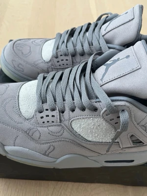 Nike Air Jordan 4 retro KAWS grå/vit - Säljer ett par Nike Air Jordan 4 KAWS i helgrå mocka med broderade detaljer och ikoniska KAWS 'XX' på hälen. Skorna har rund tå, snörning och en unik glow-in-the-dark sula. Perfekt för dig som vill sticka ut med en limiterad sneaker i premiumkänsla.   Helt ny! Storleken motsvarar exakt storlek 43. De är tyvärr helt enkelt för stora för mig. Fick den i julklapp.