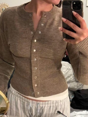 Beige ribbad kofta med knappar - Säljer en beige ribbad kofta med långa ärmar och rund hals. Koftan har silvriga knappar hela vägen framtill och är figurnära i modellen. Perfekt att slänga över en topp för en chill men ändå snygg look.