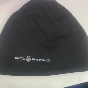 Svart Sail Racing fleecebeanie - En svart mössa från Sail Racing i mjuk fleece med diskret logga framtill. Tillverkad av återvunnen polyester och lite polyuretan för extra stretch. Enkel och stilren design som passar perfekt till kalla dagar.