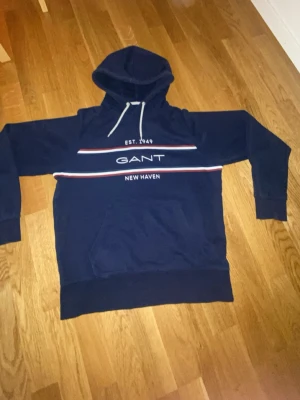 Gant hoodie  - Mörkblå GANT-hoodie med huva, ficka och tryck på bröstet. I fint skick..Köpt för 1600,inte använd så mycket bra om du gillar stilrena looks med enkla färger