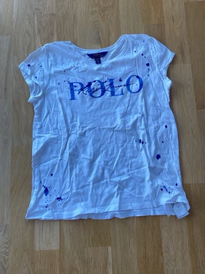 Vit POLO t-shirt med blåt tryck - Säljer en vit t-shirt från POLO Ralph Lauren med blått tryck och coola färgstänk över hela framsidan. Klassisk rund hals och korta ärmar. 